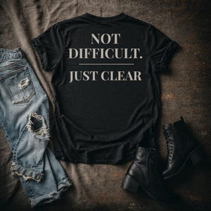 May include: Black t-shirt with the text "NOT DIFFICULT. JUST CLEAR" in white. The shirt is laid on a textured surface next to a pair of distressed blue jeans and black ankle boots.