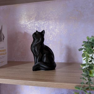 Puede incluir: Una figura de gato negro está sobre una estantería de madera, con la cabeza hacia arriba. Tres libros están a la izquierda del gato y una planta en maceta a la derecha. El fondo es una pared de color púrpura claro.