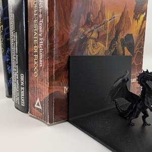 Può includere: Un fermalibri a forma di drago nero è posto davanti a una fila di libri. Il drago ha le ali spiegate ed è posizionato su una base nera ad angolo retto. I libri hanno copertine e dorsi colorati con titoli.
