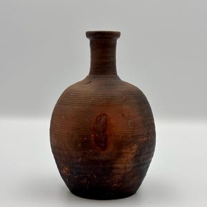 May include: A brown, handcrafted ceramic vase with a rounded body and a narrow neck. The vase has a textured surface with horizontal lines and a dark, mottled appearance. The overall design is rustic and earthy.