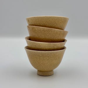 Op de afbeelding: Een stapel van vier beige keramische kommen met een craquelé glazuur. De kommen zijn op elkaar gestapeld, waarbij de onderste kom een kleine, afgeronde voet heeft. De kommen hebben een eenvoudig, elegant ontwerp.
