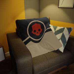 May include: A cushion featuring a red skull emblem on a navy blue backdrop, encircled by a design. It has a tan panel and is placed with a patterned cushion on a brown leather armchair.