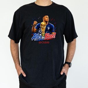 May include: Black t-shirt featuring a graphic of a soccer player holding a trophy. The design includes the text "Allez les Bleus!" and the year "2026" in red and blue. The player is wearing a blue jersey.