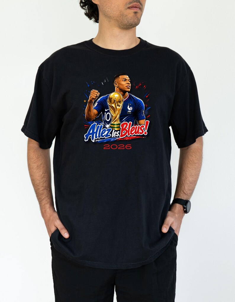 May include: Black t-shirt featuring a graphic of a soccer player holding a trophy. The design includes the text "Allez les Bleus!" and the year "2026" in red and blue. The player is wearing a blue jersey.