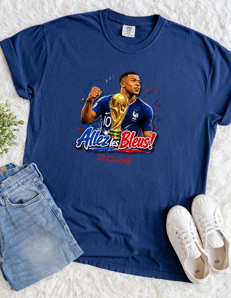 May include: Navy blue t-shirt featuring a graphic of a soccer player holding a trophy, with the text "Allez les Bleus!" and "2026". The shirt is paired with blue jeans and white sneakers.