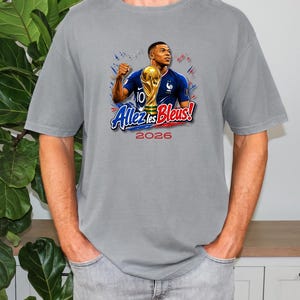 May include: Gray t-shirt featuring a graphic of a soccer player holding a trophy, with the text "Allez les Bleus!" and the year "2026". The design includes the French flag colors and a gold trophy.