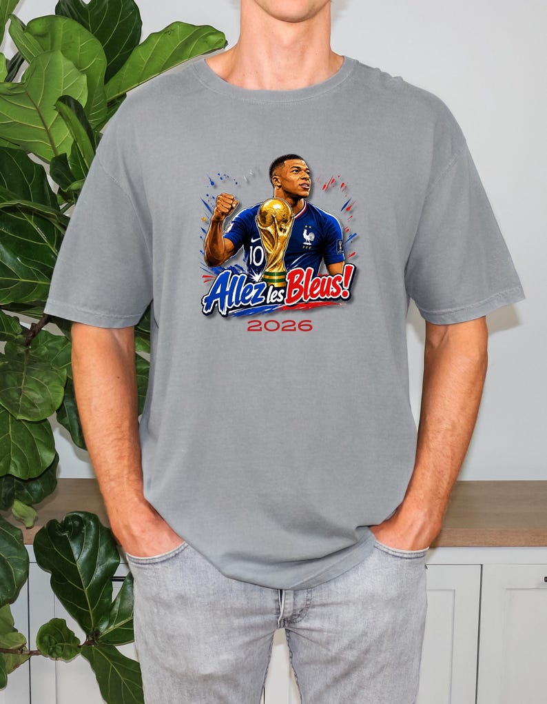 May include: Gray t-shirt featuring a graphic of a soccer player holding a trophy, with the text "Allez les Bleus!" and the year "2026". The design includes the French flag colors and a gold trophy.