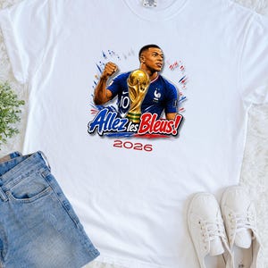 May include: White t-shirt featuring a graphic of a soccer player holding a trophy, with the text "Allez les Bleus!" and the year "2026". The player is wearing a blue jersey with the number 10. The design includes red and blue accents.