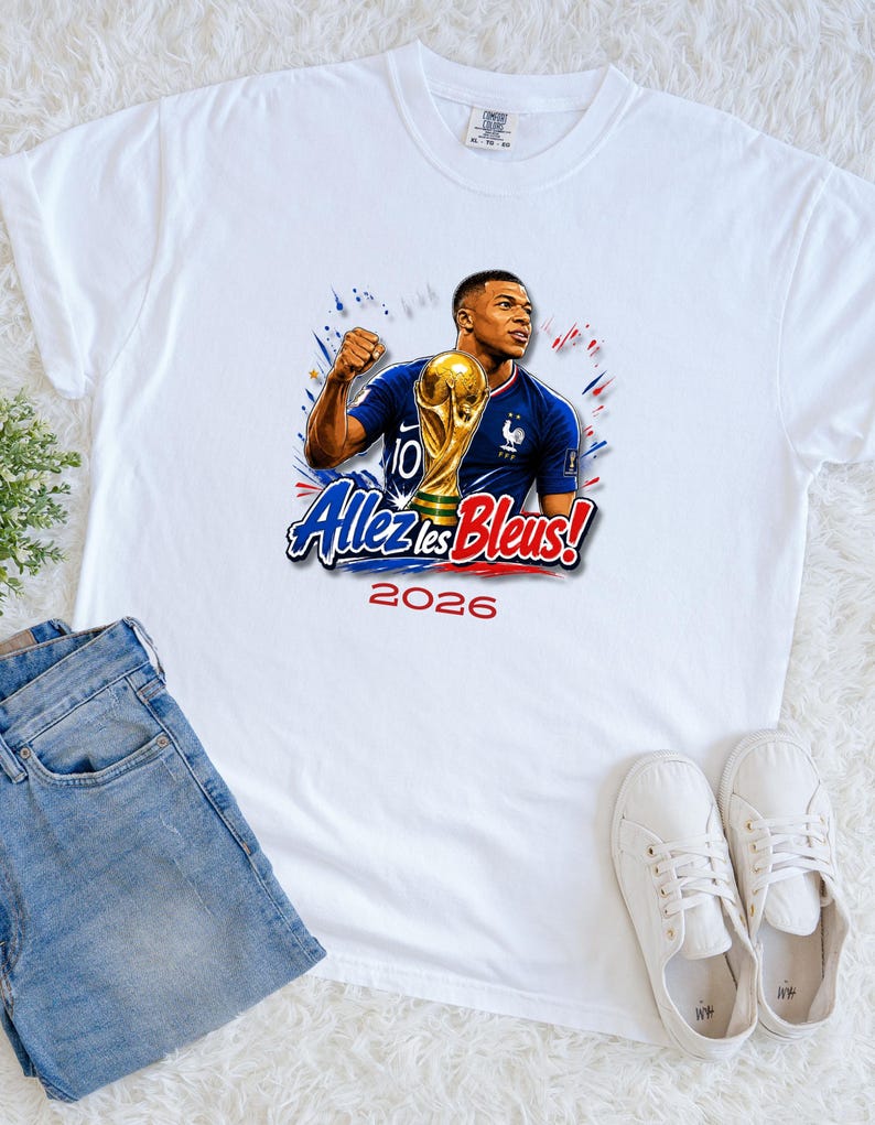 May include: White t-shirt featuring a graphic of a soccer player holding a trophy, with the text "Allez les Bleus!" and the year "2026". The player is wearing a blue jersey with the number 10. The design includes red and blue accents.