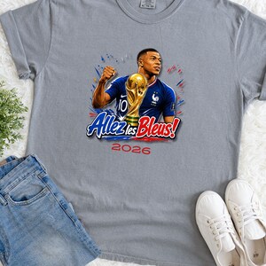 May include: Gray t-shirt featuring a graphic of a soccer player holding a trophy, with the text "Allez les Bleus!" and the year "2026". The player is wearing a blue jersey. The shirt is displayed with blue jeans and white sneakers.