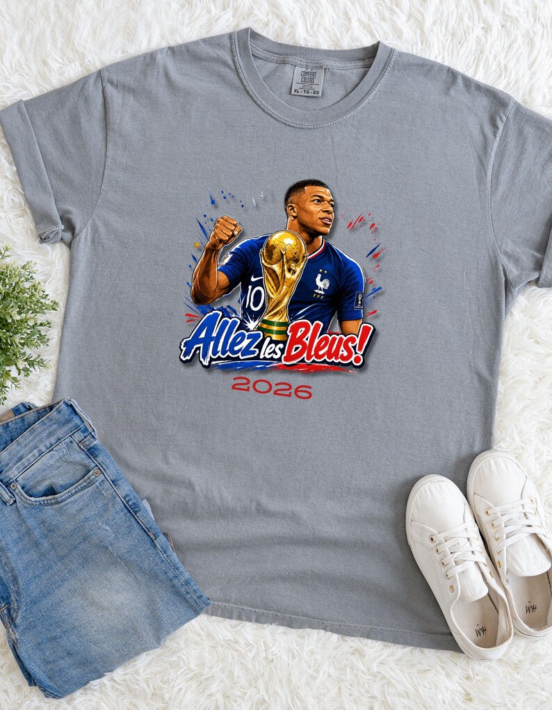 May include: Gray t-shirt featuring a graphic of a soccer player holding a trophy, with the text "Allez les Bleus!" and the year "2026". The player is wearing a blue jersey. The shirt is displayed with blue jeans and white sneakers.