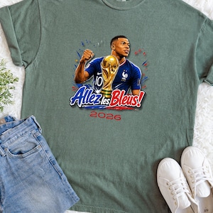May include: Sage green t-shirt featuring a graphic of a soccer player holding a trophy, with the text "Allez les Bleus!" and "2026". The shirt is paired with blue jeans and white sneakers.