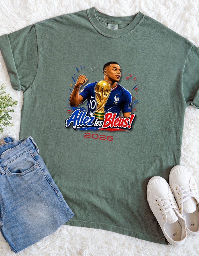 May include: Sage green t-shirt featuring a graphic of a soccer player holding a trophy, with the text "Allez les Bleus!" and "2026". The shirt is paired with blue jeans and white sneakers.