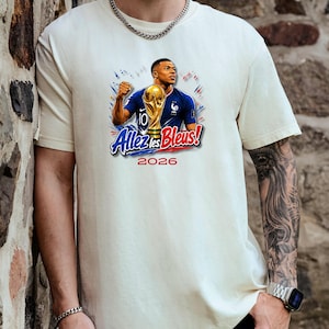 May include: Cream-colored t-shirt featuring a graphic of a soccer player holding a trophy, with the text "Allez les Bleus!" and the year "2026" in red and blue. The player is wearing a blue jersey.