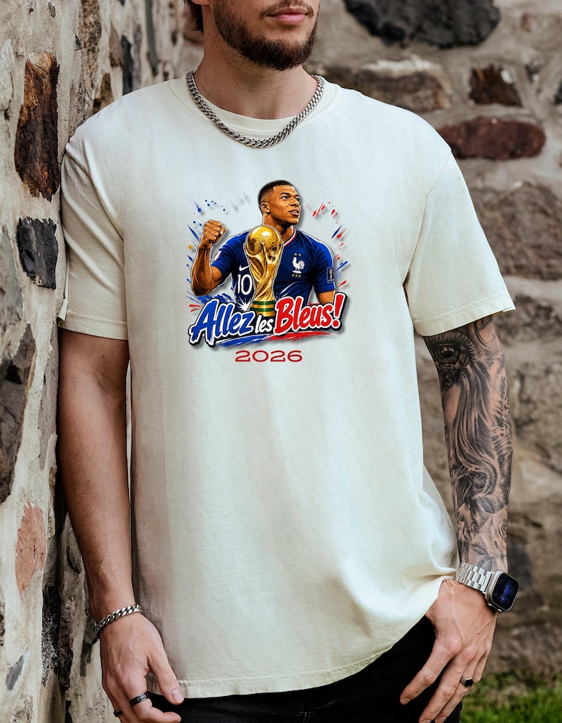 May include: Cream-colored t-shirt featuring a graphic of a soccer player holding a trophy, with the text "Allez les Bleus!" and the year "2026" in red and blue. The player is wearing a blue jersey.