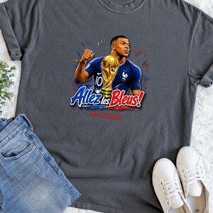 May include: Dark gray t-shirt featuring a graphic of a soccer player holding a trophy, with the text "Allez les Bleus!" and "2026". The player is wearing a blue jersey. The shirt is paired with blue jeans and white sneakers.