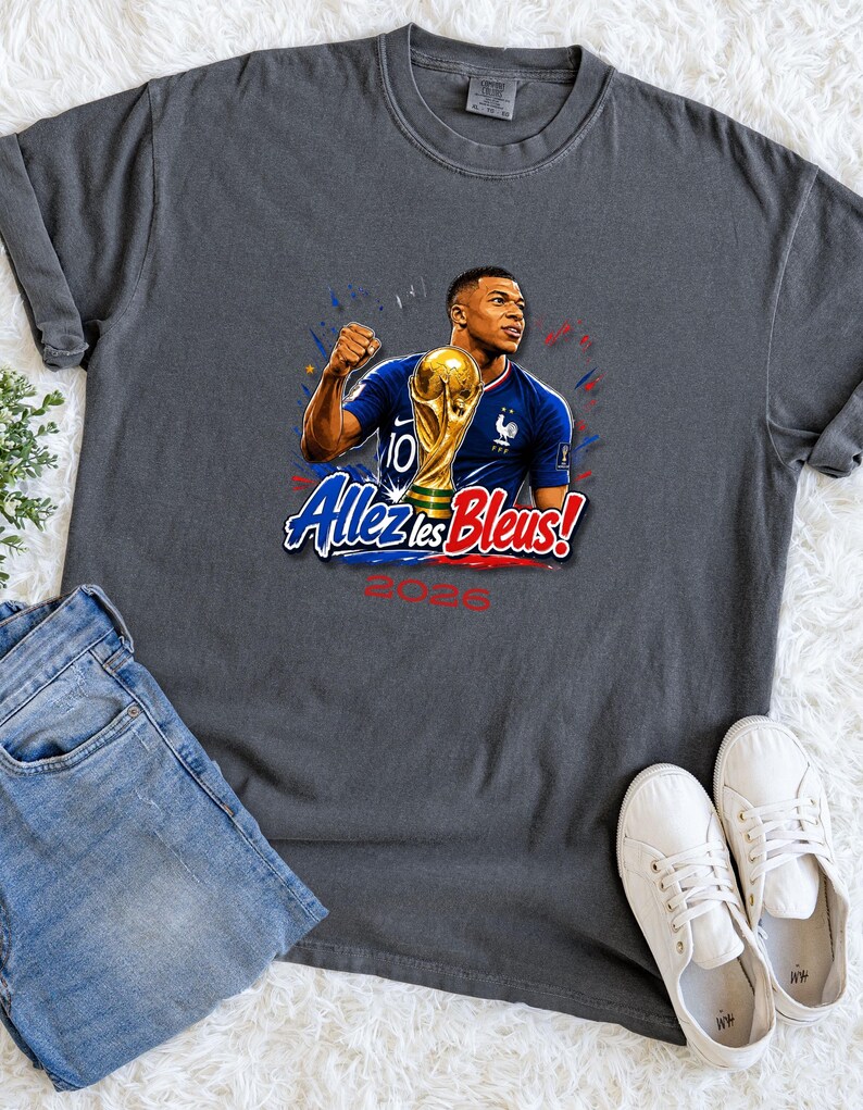May include: Dark gray t-shirt featuring a graphic of a soccer player holding a trophy, with the text "Allez les Bleus!" and "2026". The player is wearing a blue jersey. The shirt is paired with blue jeans and white sneakers.