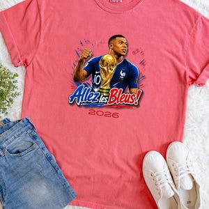 May include: A coral-colored t-shirt featuring a graphic of a soccer player holding a trophy, with the text "Allez les Bleus!" and the year "2026". The shirt is paired with blue jeans and white sneakers.