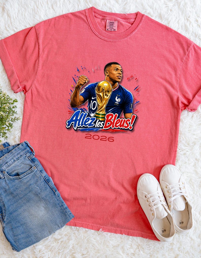 May include: A coral-colored t-shirt featuring a graphic of a soccer player holding a trophy, with the text "Allez les Bleus!" and the year "2026". The shirt is paired with blue jeans and white sneakers.