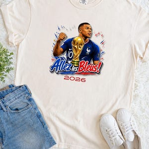 May include: Cream-colored t-shirt featuring a graphic of a soccer player holding a trophy, with the text "Allez les Bleus!" and the year "2026". The player is wearing a blue jersey. The shirt is paired with blue jeans and white sneakers.