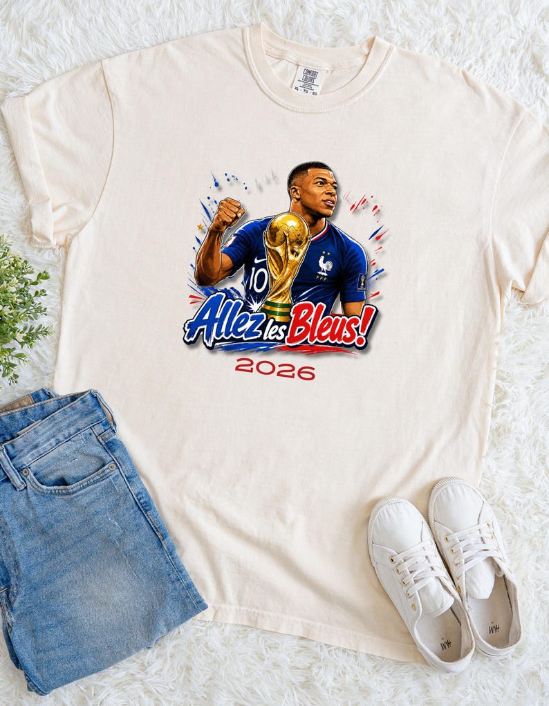 May include: Cream-colored t-shirt featuring a graphic of a soccer player holding a trophy, with the text "Allez les Bleus!" and the year "2026". The player is wearing a blue jersey. The shirt is paired with blue jeans and white sneakers.