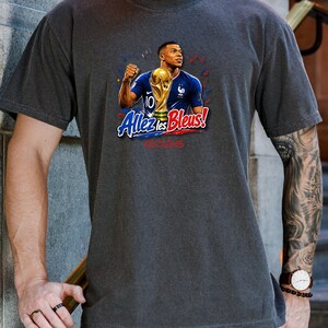 May include: Dark gray t-shirt featuring a graphic of a soccer player holding a trophy, with the text "Allez les Bleus!" and "2026". The player is wearing a blue jersey. The design includes red, white, and blue accents.