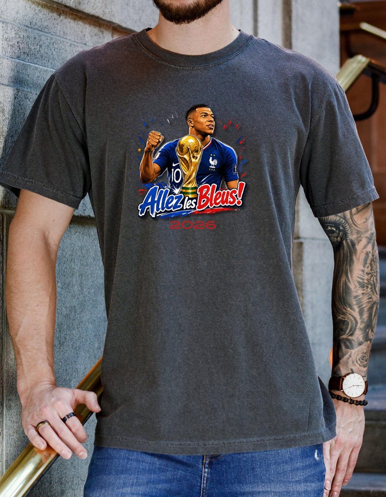 May include: Dark gray t-shirt featuring a graphic of a soccer player holding a trophy, with the text "Allez les Bleus!" and "2026". The player is wearing a blue jersey. The design includes red, white, and blue accents.