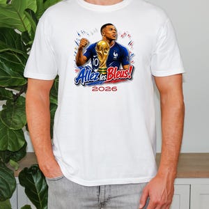 May include: White t-shirt featuring a graphic of a soccer player holding a trophy, with the text "Allez les Bleus!" and the year "2026". The design includes blue, red, and gold accents, celebrating French soccer.