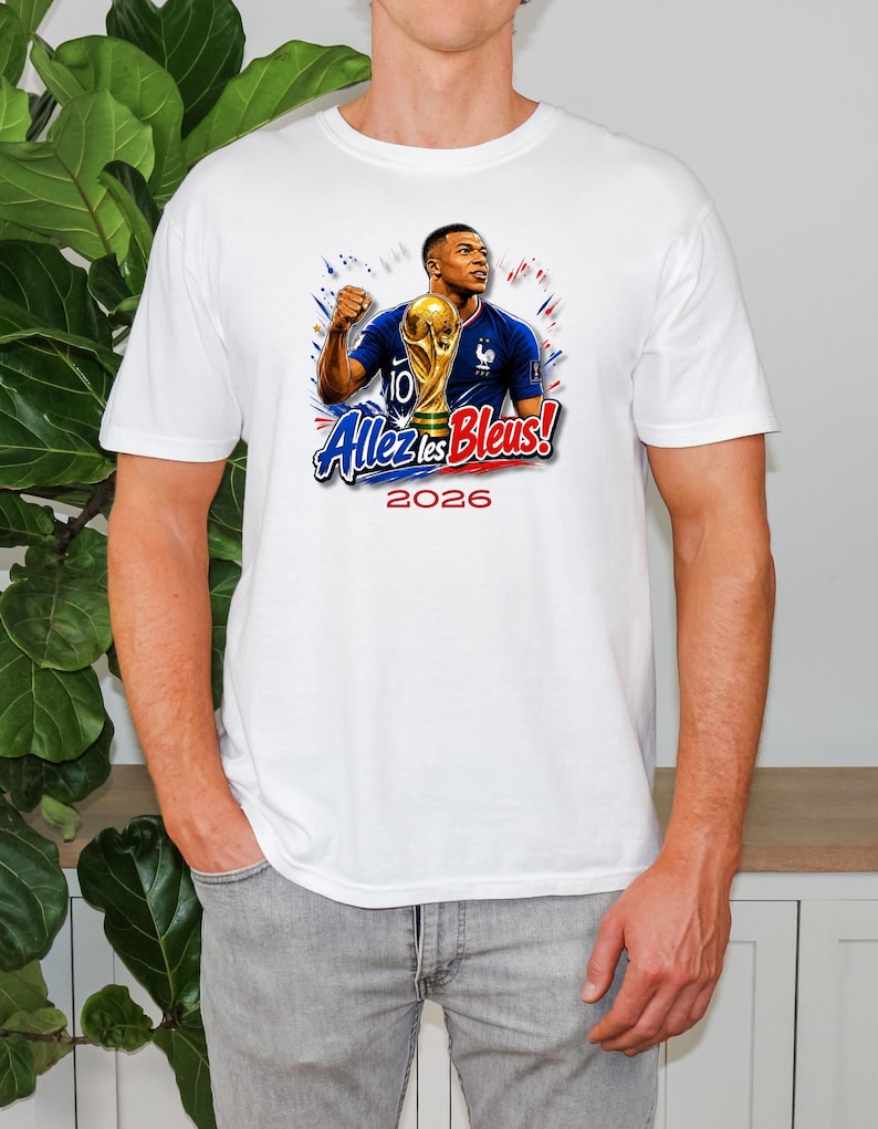 May include: White t-shirt featuring a graphic of a soccer player holding a trophy, with the text "Allez les Bleus!" and the year "2026". The design includes blue, red, and gold accents, celebrating French soccer.