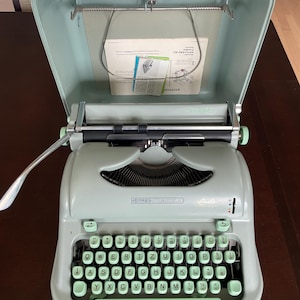 Máquina de escribir Hermes Media 3, gris verdoso, QWERTZ, en funcionamiento, con estuche y manual.