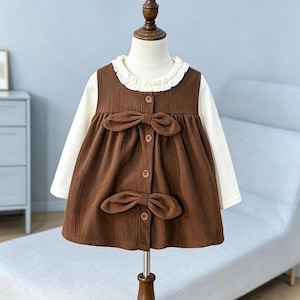 May include: A long-sleeved dress featuring a brown corduroy pinafore overlay with two decorative bows and a front button closure. The dress includes a cream-colored undershirt with a ruffled collar, creating a layered appearance on a mannequin.