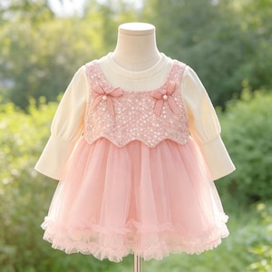 May include: A long-sleeved dress featuring a cream-coloured ribbed knit top and a pink sequined bodice overlay. The dress includes two small pink bows with pearl accents at the neckline and a layered pink tulle skirt with a ruffled hemline.