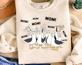 May include: Cream-colored sweatshirt featuring a cartoon illustration of seagulls with the word "MOM!" repeated around them. The seagulls are in various poses, with black and white bodies, yellow feet, and open beaks. The text "MOM!" is in a bold, black font.