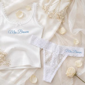 May include: A white ribbed tank top with lace trim and a small bow, featuring the embroidered text Mrs. Brown in light blue. A matching white lace thong with a small bow and the embroidered text Mrs. Brown in light blue is positioned nearby.