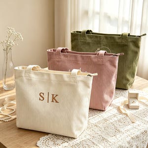 Op de afbeelding: Drie corduroy tote bags in crème, oudroze en olijfgroen. De crèmekleurige tas is voorzien van de geborduurde letters S | K. Elke tas heeft twee handvatten, een ritssluiting en een rechthoekige vorm, gerangschikt in een rij op een tafel.