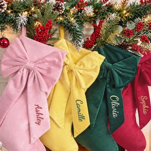 May include: Four velvet Christmas stockings in light pink, yellow, dark green, and red. Each stocking features a large decorative bow on the front and is personalized with embroidered names: Ashley, Camille, Olivia, and Sophia in script font.