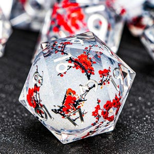 May include: A clear, multi-sided die with a floral design. The die features red flowers and black branches encased in a clear resin. The numbers are silver. The background is a glittery white.