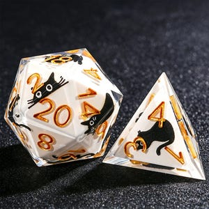 May include: Two polyhedral dice, a 20-sided die and a 4-sided die, are shown. The dice are clear with white interiors. Black cat illustrations and gold numbers are visible on the faces. The dice are designed for tabletop games.