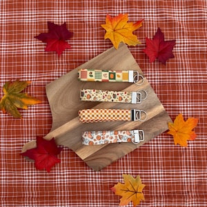 May include: Four fabric wristlet keychains featuring autumn-themed patterns including pumpkins, leaves, and checkered designs. Each keychain has a silver-toned metal hardware clasp and a circular split ring attached to one end for holding keys.