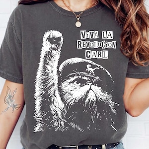 May include: A charcoal grey short-sleeve t-shirt featuring a white graphic print of a cat wearing a beret with a small dinosaur icon. The cat has one paw raised, and the text VIVA LA REVOLUCION CARL is printed in white above the cat's head.
