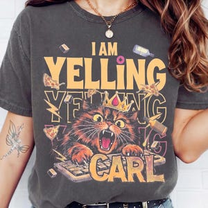 May include: A charcoal grey short-sleeve t-shirt featuring a graphic of a cat wearing a crown while typing on a keyboard. The text reads I AM YELLING YELLING YELLING CARL, surrounded by illustrations of pizza slices, bottles, and lightning bolts.