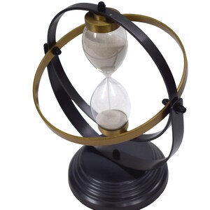 May include: A rotating hourglass featuring a clear glass bulb filled with light-colored sand, encased within a dual-ring frame of matte black and metallic gold. The entire structure is supported by a solid, circular, tiered matte black base.