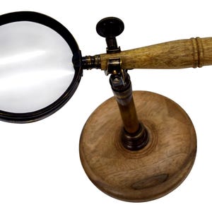 May include: A magnifying glass featuring a circular lens with a black frame, attached to a horizontal wooden handle. The assembly is mounted on a vertical brass-toned metal post connected to a circular wooden base, allowing for adjustable positioning.