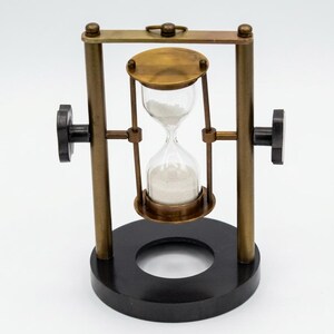 May include: A rotating hourglass featuring a clear glass bulb filled with white sand, held within a brass frame. The frame is supported by two vertical brass pillars attached to a circular black base, with black adjustment knobs on each side.