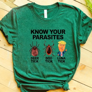 以下が含まれることがあります： ヘザーグリーンの半袖Tシャツ。黒い文字で「KNOW YOUR PARASITES」と書かれ、その下に3つのイラストが並んでいます。シカダニ、イヌダニ、そして「LUNA TICK」とラベル付けされた似顔絵が描かれ、それぞれのイラストの下に黒い文字でラベルが添えられています。