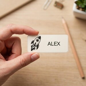 May include: A rectangular white name label featuring a black line art illustration of a rocket ship on the left side and the name ALEX printed in bold black capital letters on the right side of the smooth, matte surface of the sticker.