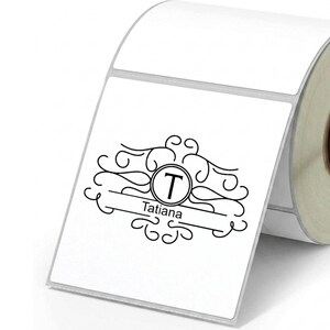 Peut inclure: Un rouleau d'étiquettes adhésives rectangulaires blanches avec un motif imprimé noir. Le design comprend un cercle central contenant la lettre T, entouré de fioritures décoratives et d'une ligne horizontale avec le nom Tatiana imprimé dessous.