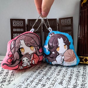 May include: Two plush charms featuring anime-style characters with long brown hair and amber eyes. One charm has a pink background with a character holding a small figure, while the other has a blue background with a character holding a round tan object.