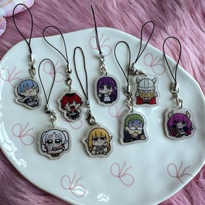May include: A collection of eight clear acrylic charms featuring colourful chibi-style anime characters. Each charm is attached to a black cord loop with a silver-toned metal clasp. The characters include various figures with distinct hair colours and outfits.