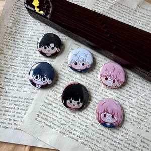 May include: Six circular pin-back buttons featuring stylized chibi-style character illustrations. The buttons display various hair colors including black, white, and pink, with expressive facial features. The pins are arranged on a page of text.